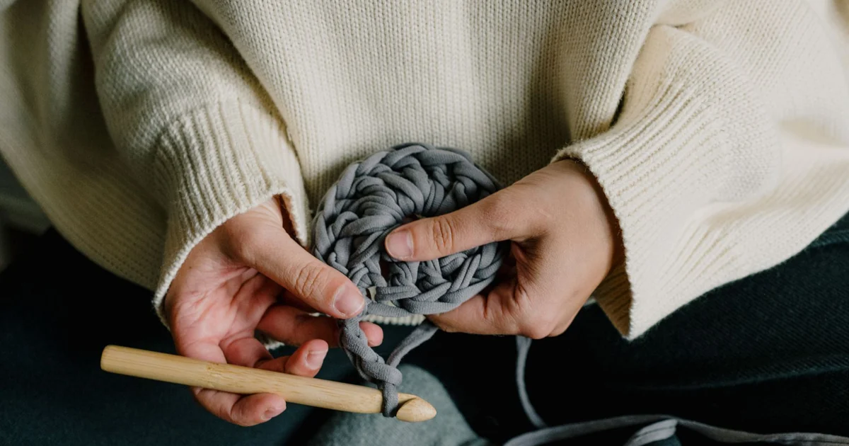 Hands crocheting a gray-blue piece beside yarn.