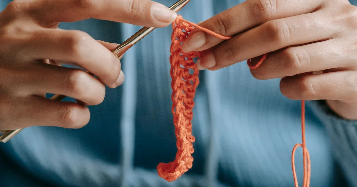 Hands working coral yarn across a narrow crochet piece.