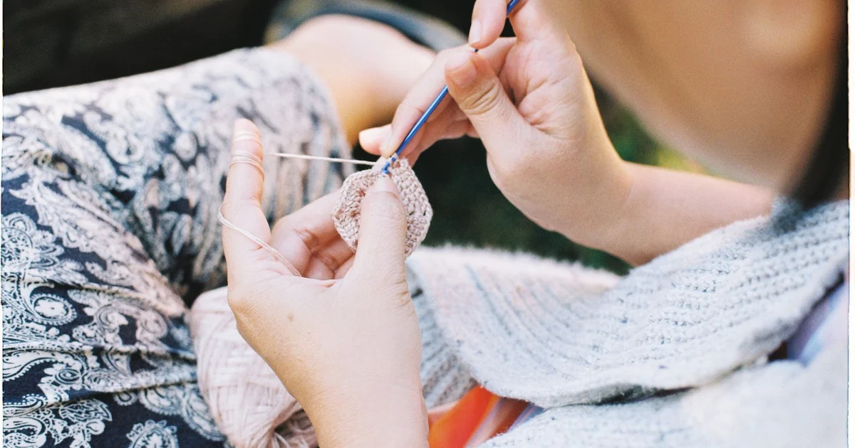 Hands working a blue crochet hook along the edge of a light crochet fabric.
