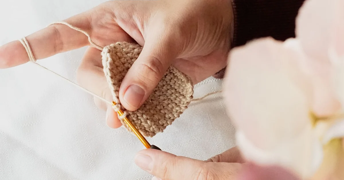 Hands using a gold crochet hook on a brown crochet swatch.