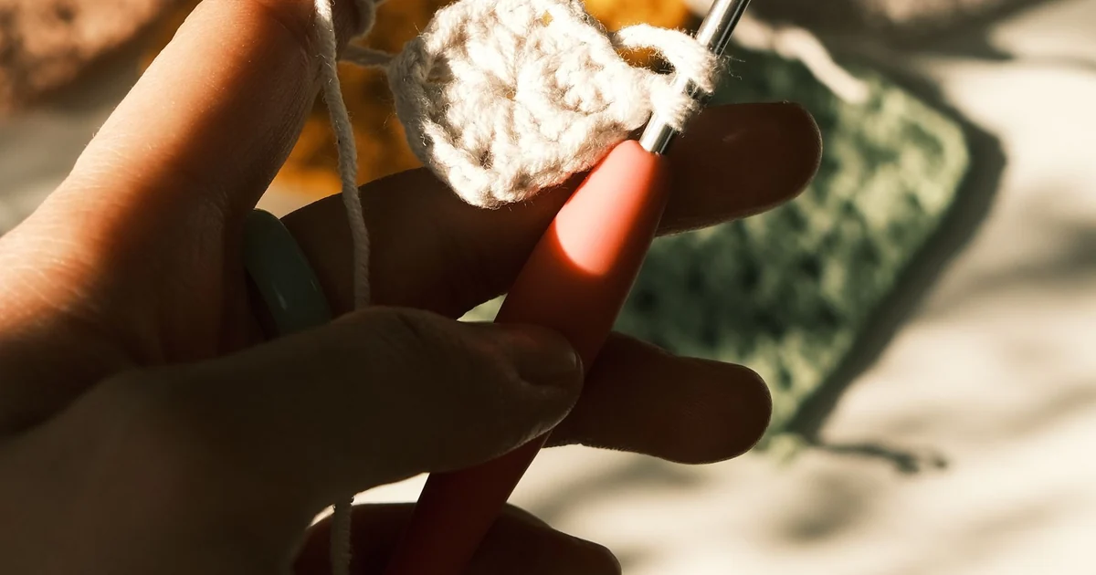 Hands holding a pink crochet hook and white yarn against a dark background.