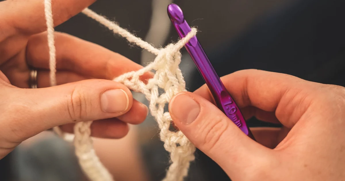 Purple hook and hands working yarn beside stored crochet supplies.